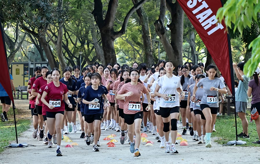 A large group of students in running attire set off from the start line of an outdoor race along a tree-lined path.